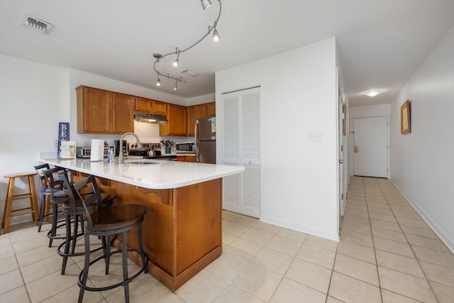 kitchen featuring stainless steel appliances, light countertops, brown cabinetry, under cabinet range hood, and a peninsula