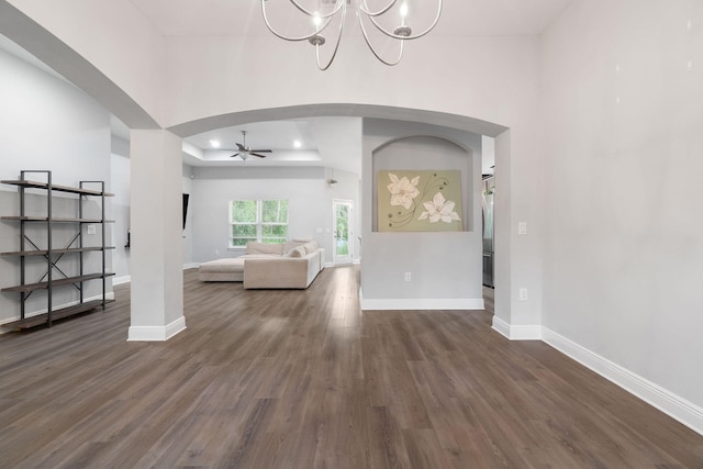 unfurnished living room featuring arched walkways, ceiling fan with notable chandelier, baseboards, dark wood-style floors, and a raised ceiling