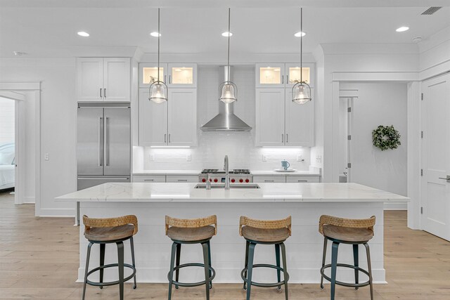 kitchen featuring wall chimney range hood, light hardwood / wood-style flooring, an island with sink, built in fridge, and sink