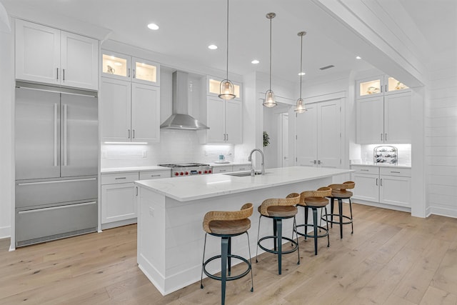 kitchen featuring wall chimney exhaust hood, built in refrigerator, a center island with sink, and white cabinets