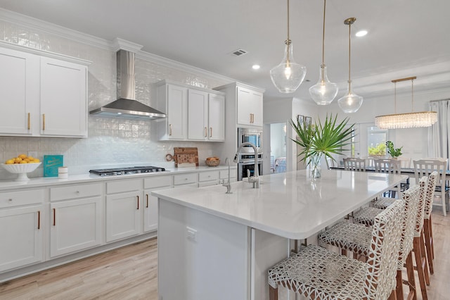 kitchen featuring wall chimney range hood, decorative backsplash, light wood-type flooring, hanging light fixtures, and stainless steel appliances