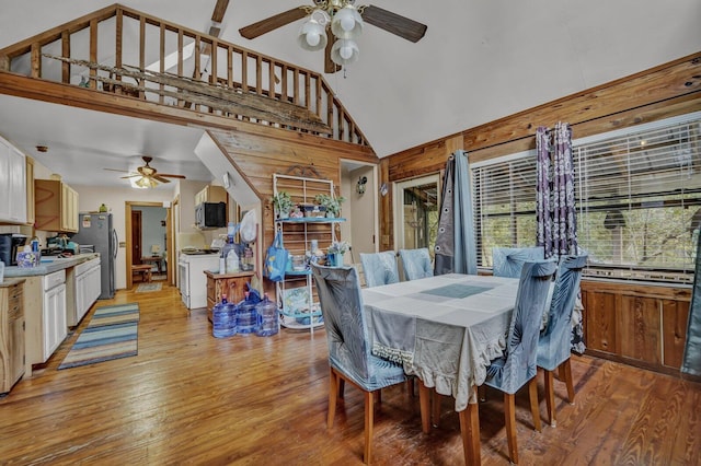 dining space featuring ceiling fan, high vaulted ceiling, light hardwood / wood-style flooring, and wooden walls