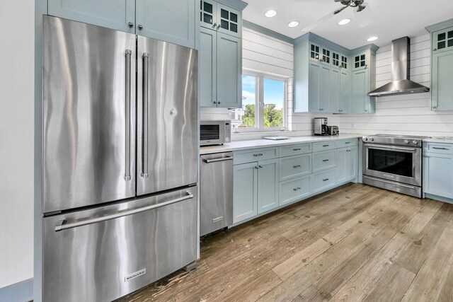 kitchen with blue cabinetry, tasteful backsplash, wall chimney exhaust hood, appliances with stainless steel finishes, and light hardwood / wood-style floors