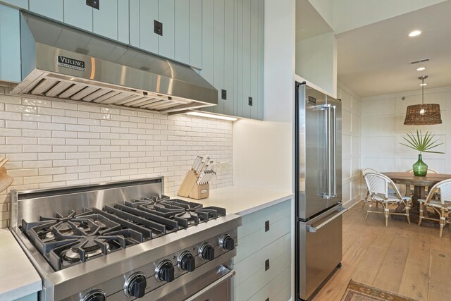 kitchen with backsplash, light wood-type flooring, wall chimney exhaust hood, high quality appliances, and hanging light fixtures