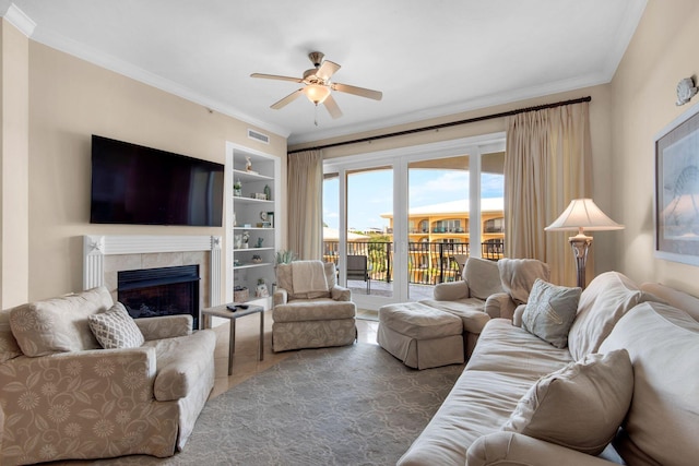 living room featuring ceiling fan, a tiled fireplace, ornamental molding, and built in shelves