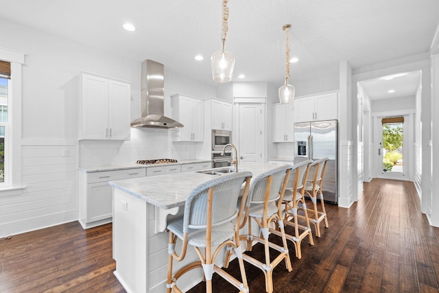 kitchen with dark hardwood / wood-style floors, wall chimney exhaust hood, decorative light fixtures, a center island with sink, and stainless steel appliances