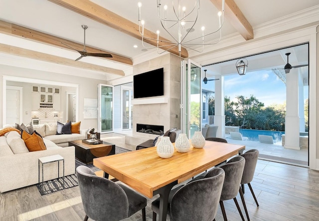 dining room with ceiling fan with notable chandelier, a tiled fireplace, beamed ceiling, light hardwood / wood-style flooring, and ornamental molding