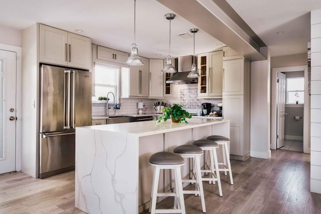 kitchen with a sink, light wood-style floors, high end fridge, wall chimney range hood, and backsplash