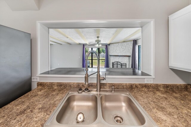 interior details featuring ceiling fan, a stone fireplace, white cabinets, beam ceiling, and sink