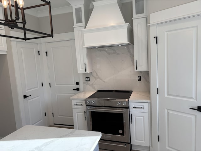 kitchen featuring backsplash, premium range hood, stainless steel electric stove, a notable chandelier, and white cabinets