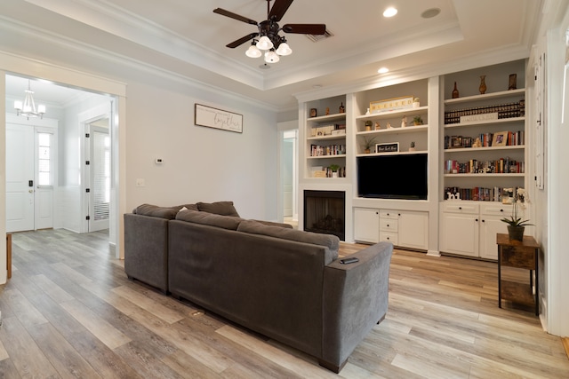 living room featuring light hardwood / wood-style flooring, a tray ceiling, ornamental molding, built in shelves, and ceiling fan with notable chandelier