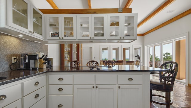 kitchen with white cabinetry, a kitchen breakfast bar, backsplash, dark stone countertops, and glass insert cabinets