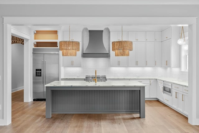 kitchen with custom exhaust hood, white cabinets, built in appliances, an island with sink, and light stone counters