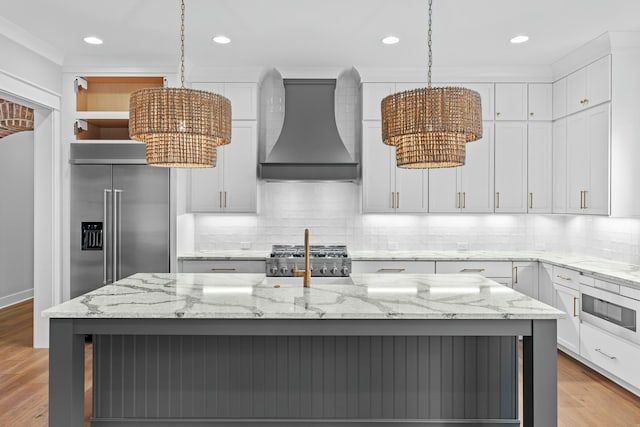 kitchen featuring high end refrigerator, premium range hood, light stone counters, decorative light fixtures, and an island with sink