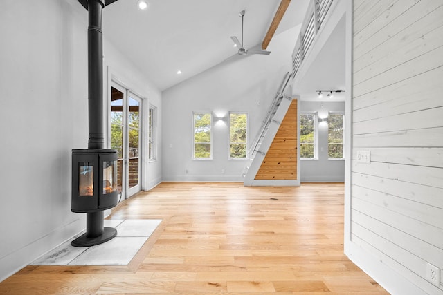 unfurnished living room featuring high vaulted ceiling, wood walls, a wood stove, ceiling fan, and light hardwood / wood-style floors