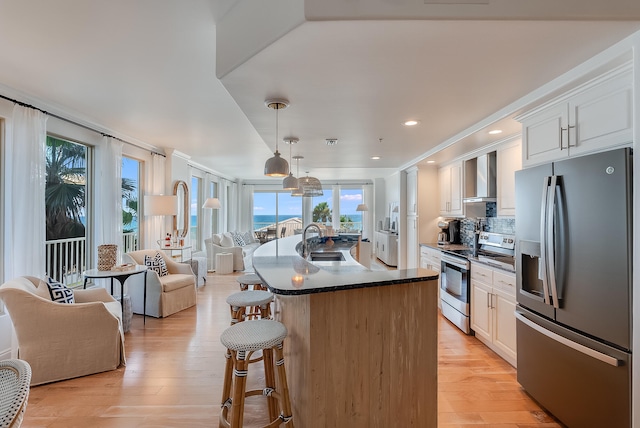 kitchen featuring wall chimney range hood, appliances with stainless steel finishes, a center island with sink, and white cabinetry