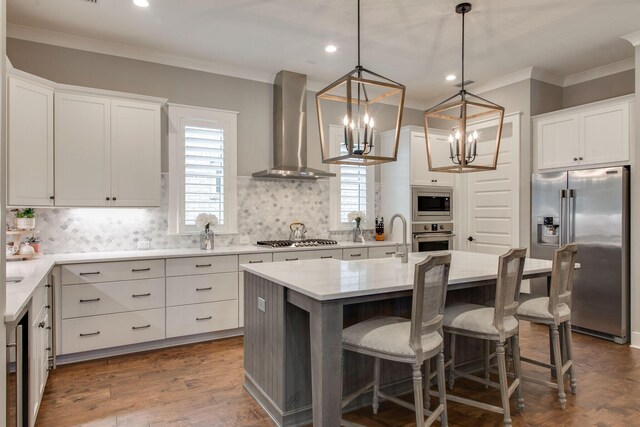 kitchen with wall chimney exhaust hood, stainless steel appliances, decorative light fixtures, a center island with sink, and white cabinets