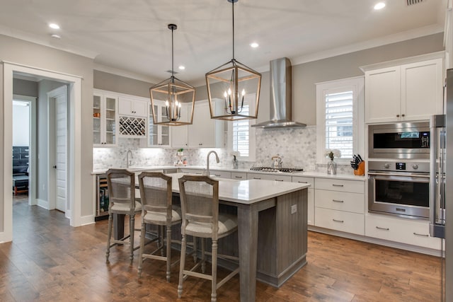 kitchen featuring pendant lighting, wall chimney exhaust hood, an island with sink, appliances with stainless steel finishes, and white cabinetry