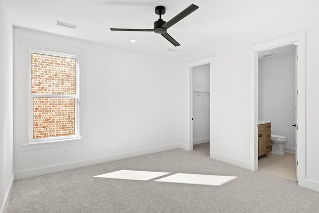 unfurnished bedroom featuring light carpet, a spacious closet, ensuite bath, and ceiling fan