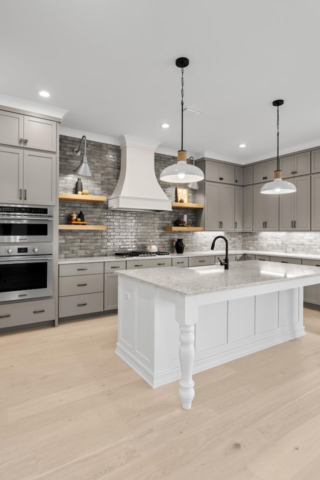 kitchen with light stone countertops, custom range hood, a kitchen island with sink, sink, and light hardwood / wood-style floors