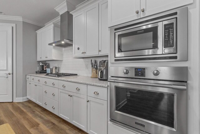 kitchen with wall chimney exhaust hood, crown molding, stainless steel appliances, white cabinetry, and hardwood / wood-style flooring