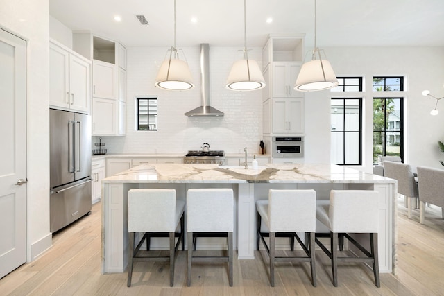 kitchen with appliances with stainless steel finishes, wall chimney range hood, a kitchen island with sink, and white cabinets