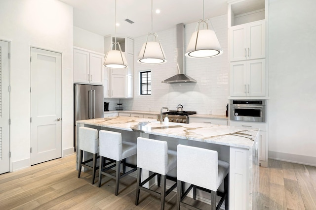 kitchen with a kitchen island with sink, oven, white cabinets, pendant lighting, and wall chimney range hood