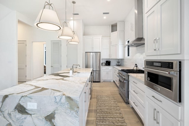 kitchen featuring appliances with stainless steel finishes, wall chimney exhaust hood, a center island with sink, and white cabinets