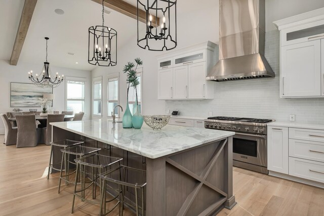 kitchen with high end stove, white cabinetry, wall chimney exhaust hood, and a kitchen island with sink