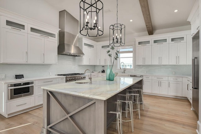 kitchen with white cabinets, light hardwood / wood-style floors, a notable chandelier, wall chimney exhaust hood, and a kitchen island with sink