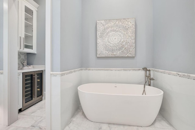 full bathroom featuring a freestanding tub, beverage cooler, marble finish floor, and wainscoting