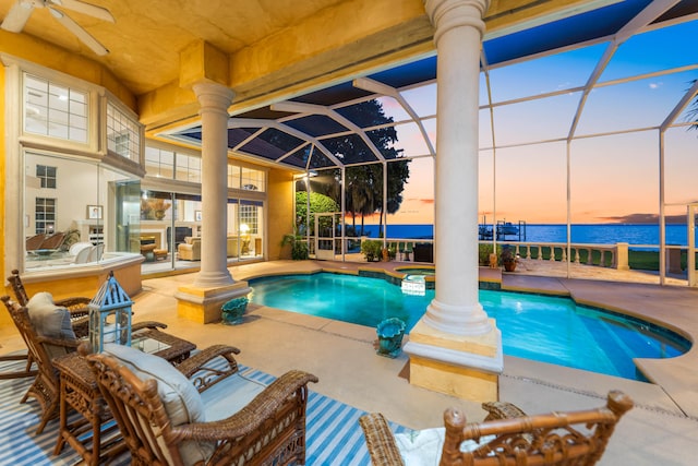 pool at dusk featuring a water view, ceiling fan, a patio area, and glass enclosure