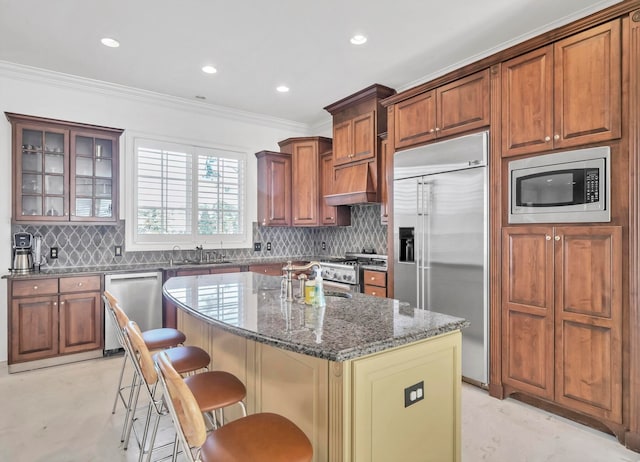 kitchen with dark stone countertops, ornamental molding, built in appliances, a breakfast bar area, and a center island with sink