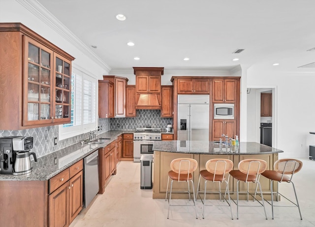 kitchen with dark stone counters, crown molding, built in appliances, an island with sink, and a breakfast bar area