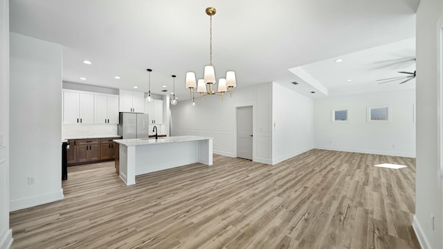 kitchen featuring hanging light fixtures, light hardwood / wood-style flooring, stainless steel fridge, a kitchen island with sink, and ceiling fan with notable chandelier