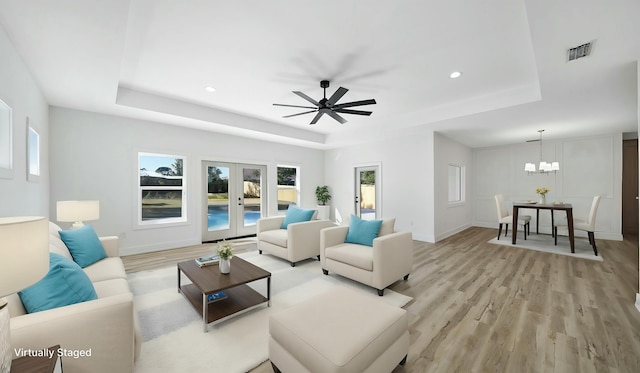 living room with french doors, ceiling fan with notable chandelier, a tray ceiling, and light hardwood / wood-style flooring