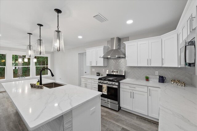 kitchen featuring a kitchen island with sink, wall chimney exhaust hood, stainless steel gas stove, decorative light fixtures, and white cabinetry