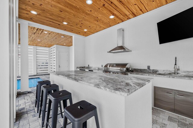 kitchen featuring light stone counters, exhaust hood, wooden ceiling, a kitchen bar, and sink