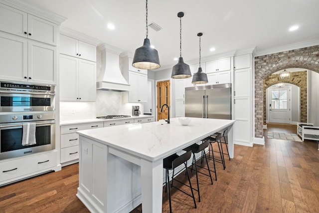 kitchen with appliances with stainless steel finishes, premium range hood, a kitchen island with sink, and white cabinetry