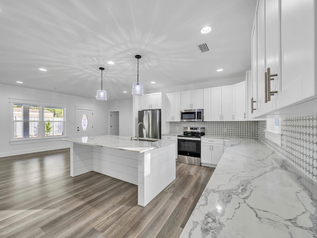 kitchen with an island with sink, white cabinets, pendant lighting, stainless steel appliances, and backsplash