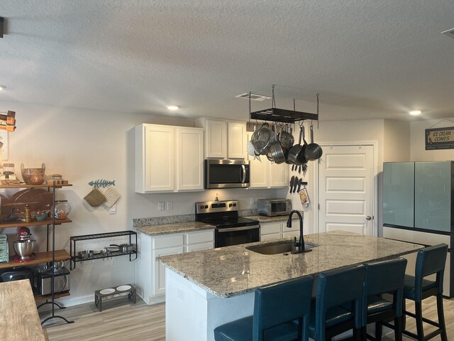 kitchen with an island with sink, sink, white cabinetry, stainless steel appliances, and light wood-type flooring