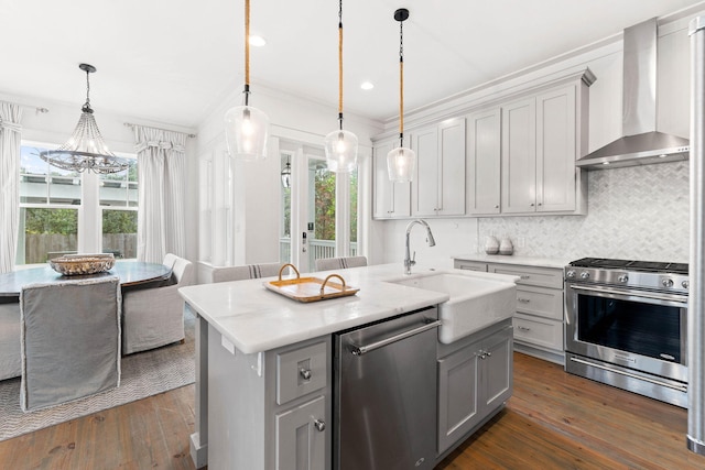 kitchen featuring wall chimney exhaust hood, an island with sink, stainless steel appliances, pendant lighting, and gray cabinets