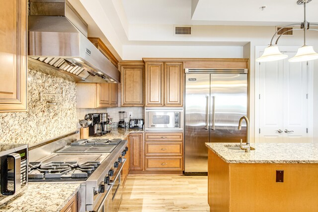 kitchen with light stone countertops, wall chimney range hood, built in appliances, decorative light fixtures, and light hardwood / wood-style floors