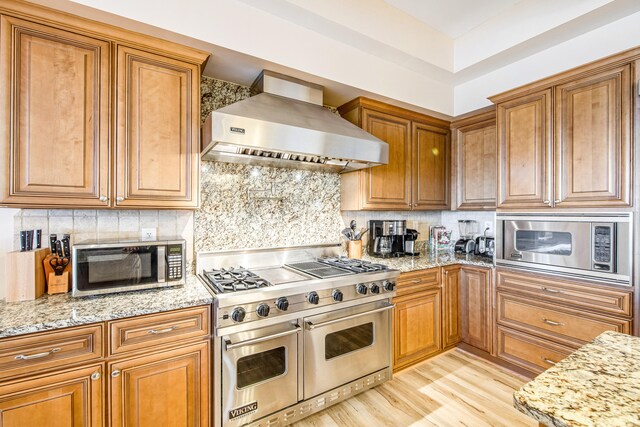 kitchen with wall chimney exhaust hood, light stone counters, light hardwood / wood-style floors, decorative backsplash, and appliances with stainless steel finishes