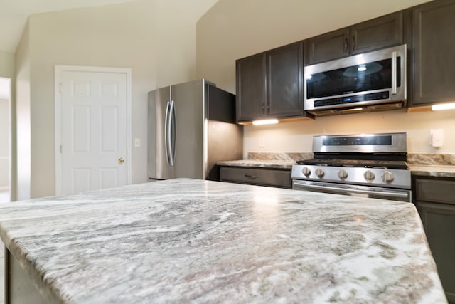 kitchen with stainless steel appliances, dark brown cabinetry, light stone counters, a kitchen island, and lofted ceiling