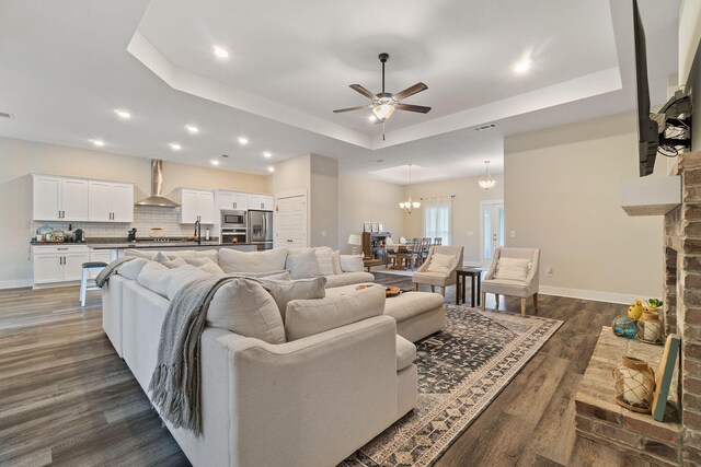 living room with ceiling fan with notable chandelier, a tray ceiling, and dark wood-type flooring