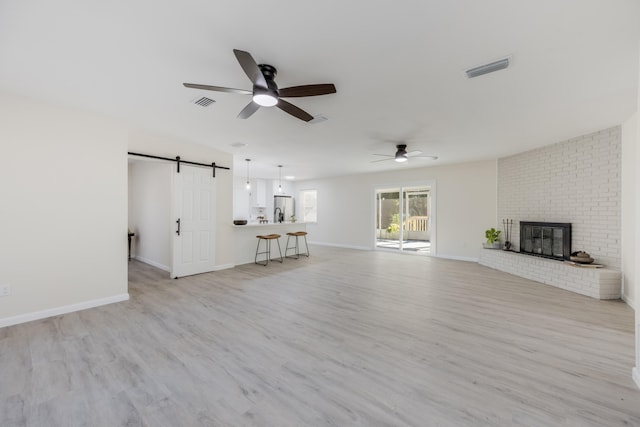 unfurnished living room with a brick fireplace, light wood-type flooring, a barn door, and ceiling fan