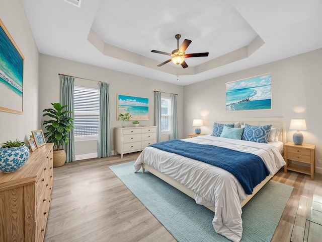 bedroom with multiple windows, a tray ceiling, light wood-type flooring, and ceiling fan