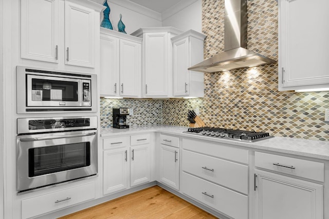 kitchen with white cabinets, tasteful backsplash, wall chimney range hood, appliances with stainless steel finishes, and light wood-type flooring