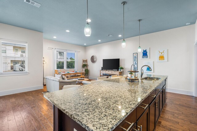 kitchen with an island with sink, light stone countertops, dark wood-type flooring, dark brown cabinetry, and sink